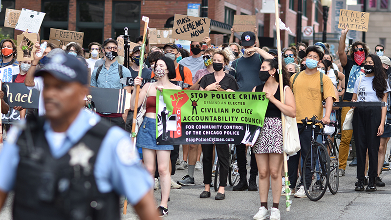 Protesters marching down a Chicago city street holding signs for police accountability and community oversight behind an officer.