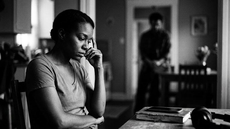 Contemplative Woman at Table (contemplative-woman.jpg) Alt Text: Anxious African American woman seated with hand on forehead while a blurred figure stands in the background.