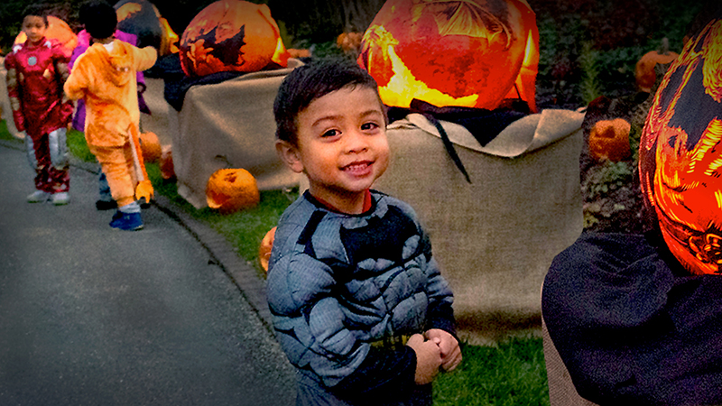 A smiling young boy dressed in a Batman costume stands on an outdoor pathway lined with glowing, hand-carved pumpkins alongside other children in costumes.