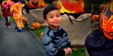 A smiling young boy dressed in a Batman costume stands on an outdoor pathway lined with glowing, hand-carved pumpkins alongside other children in costumes.