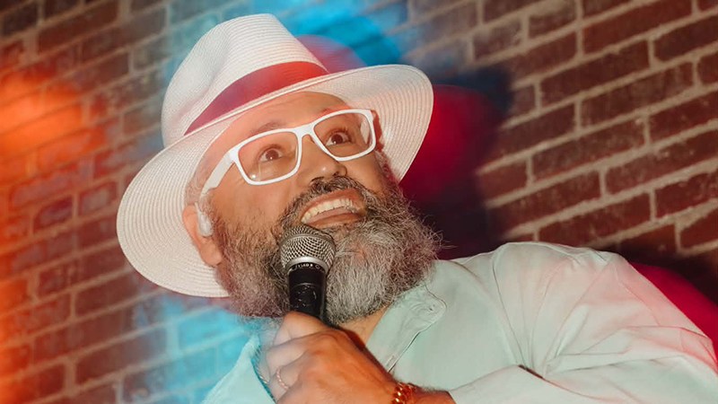 Stand-up comedian Tio Pepe Cabrera smiling into a handheld microphone against an exposed brick wall during a live stage routine.